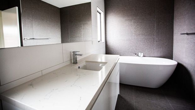 Contemporary bathroom with a long white engineered-stone vanity top, integrated rectangular basins, pale cabinetry, large mirror, chrome tapware, and a freestanding white oval bathtub. Dark textured wall and floor finishes create a strong contrast, with natural light entering through a narrow window.