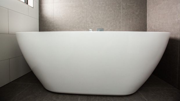 Close-up view of a contemporary bathroom dominated by a large freestanding white oval bathtub. The tub sits on dark grey tiled flooring, with large grey wall tiles behind it and lighter rectangular wall tiles to the left. Minimal fixtures and high-level glazing contribute to a clean, understated design.