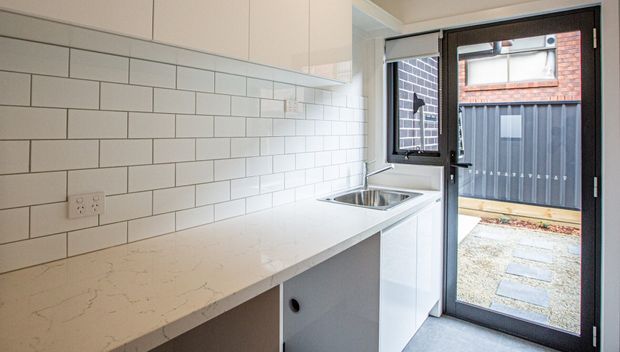 Compact contemporary kitchen or kitchenette with white flat-panel cabinetry, a pale engineered-stone benchtop, and a white rectangular subway-tile splashback. A stainless-steel sink sits beneath a window, with a glazed external door opening to a small paved courtyard or service area. The space has a clean, bright, neutral fit-out.
