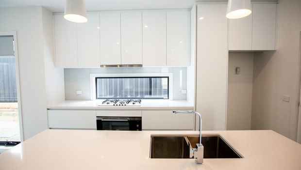Bright, compact contemporary kitchen with glossy white overhead and base cabinetry, a pale stone island benchtop, integrated sink and chrome mixer tap, gas cooktop, and built-in oven. A horizontal window provides natural light behind the cooking zone, with simple white pendant lights overhead.
