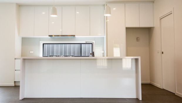 Bright, minimalist contemporary kitchen with glossy white full-height cabinetry, a large white island with an integrated raised benchtop, stainless steel rangehood and splashback area, pendant lights, and dark flooring. A doorway and adjacent cabinetry are visible to the right.
