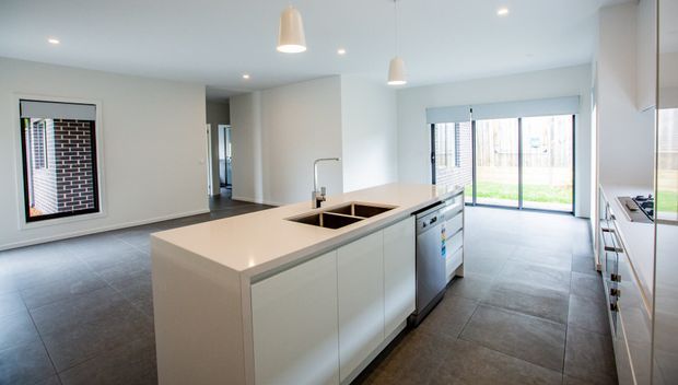 Bright, contemporary open-plan kitchen with a large central island, integrated sink, dishwasher, white cabinetry, and pale stone benchtops. Large sliding glass doors open to a small lawn or outdoor area, while dark large-format floor tiles and recessed ceiling lights provide contrast. A separate living or dining zone is visible to the left.