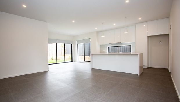 Bright open-plan kitchen and living area with glossy white built-in cabinetry, a central island, large black-framed glazed doors and windows, recessed ceiling lighting, and large dark grey tiled flooring. The space is unfurnished and finished in a clean contemporary style.