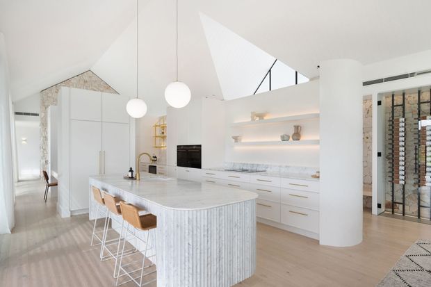 Bright contemporary white kitchen with a large curved island featuring vertically fluted detailing and seating for four. The space includes white cabinetry, pale timber flooring, marble-look benchtops and backsplash, pendant globe lights, open display shelving, a cylindrical structural column, and a high angular skylight or clerestory opening. A wine storage area is visible behind glazed doors.