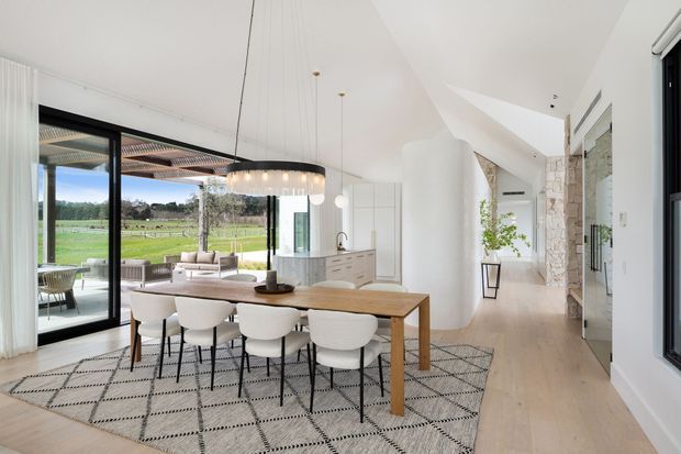 Bright open-plan dining and living space with a long timber dining table and six upholstered chairs on a patterned rug. Floor-to-ceiling black-framed glazing opens to an outdoor entertaining area and lawn, while a white kitchen with an island, pendant lights, a sculptural cylindrical column, and a stone-clad hallway wall extend through the background. The interior is airy and contemporary with pale timber flooring and white finishes.