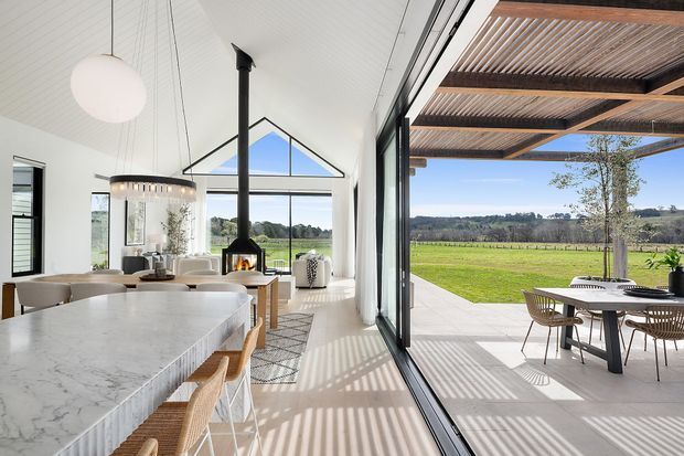 Bright, open-plan contemporary kitchen and dining area opening through full-height glazed sliding doors to a covered outdoor terrace. The interior features a large white marble-look island, timber stools, a pitched white-lined ceiling, suspended pendant lighting, and a black freestanding fireplace. The terrace has a timber slatted pergola, outdoor dining furniture, and views across a green rural landscape.