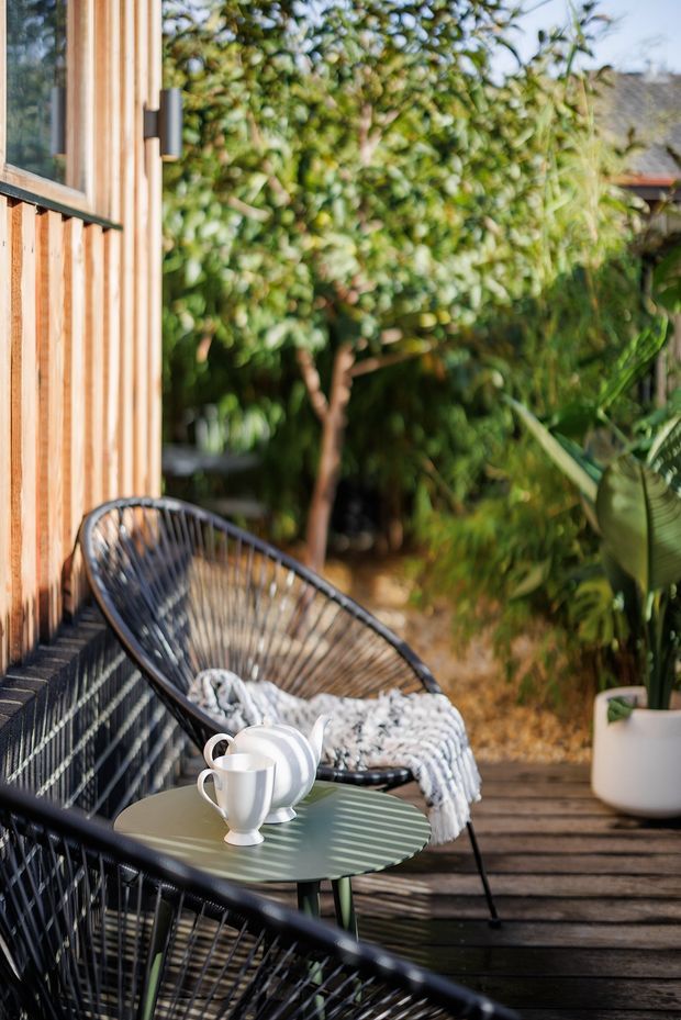 A sunlit outdoor deck beside a timber-clad wall, furnished with black woven lounge chairs, a small green round table, white crockery, and a patterned throw. Dense greenery and potted tropical planting create a private garden setting. The image has a relaxed, natural, resort-like atmosphere.