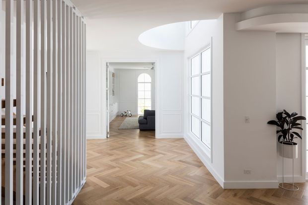 Bright contemporary interior hallway or entry area with white walls, large gridded windows, a curved ceiling opening, vertical white slatted screening, and light timber herringbone flooring. A living room is visible through the open doorway in the background.