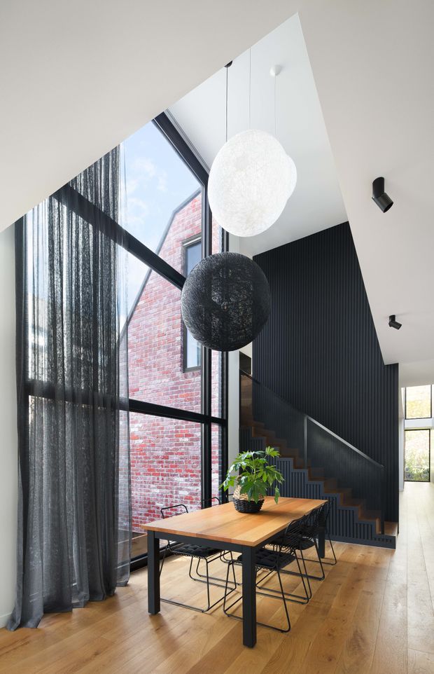 A contemporary double-height dining area with a timber dining table and black wire-frame chairs, positioned beside expansive black-framed glazing with sheer curtains. Two oversized spherical pendant lights hang above the table. A dark vertically ribbed feature wall forms the backdrop to a timber staircase, while an exposed red-brick wall is visible through the glazing. The room has warm timber flooring, crisp white walls and ceiling, and strong natural light.