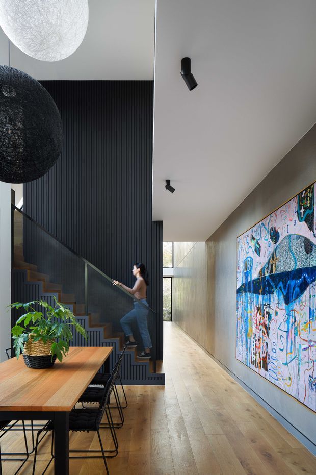 A contemporary open-plan interior combining a dining area, stairway, and long hallway. The space features light timber flooring, a timber dining table with black metal-framed chairs, a dramatic black vertically slatted stair wall, glass stair balustrade, white ceilings with black cylindrical track spotlights, and a large colourful abstract artwork along the hallway wall. Oversized white and black pendant lights and indoor planting add visual interest.