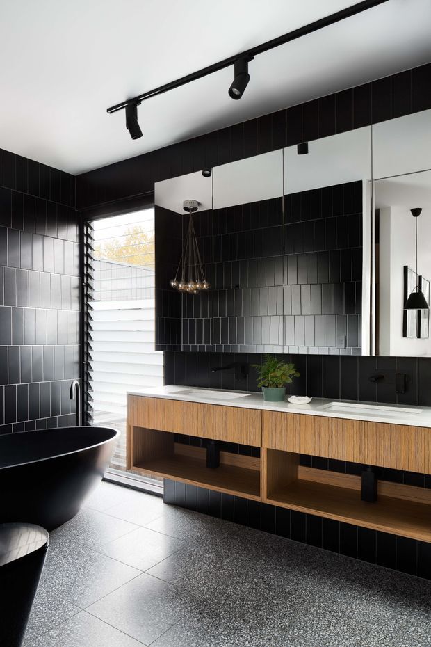 A dramatic contemporary bathroom featuring a freestanding matte-black bathtub, large mirrored wall, twin vanity with warm timber cabinetry and white benchtop, and extensive vertically stacked black wall tiles. The room has a dark terrazzo-like tiled floor, black track lighting, a sculptural pendant, louvred window screening, and a clean monochromatic palette softened by timber and a small plant.