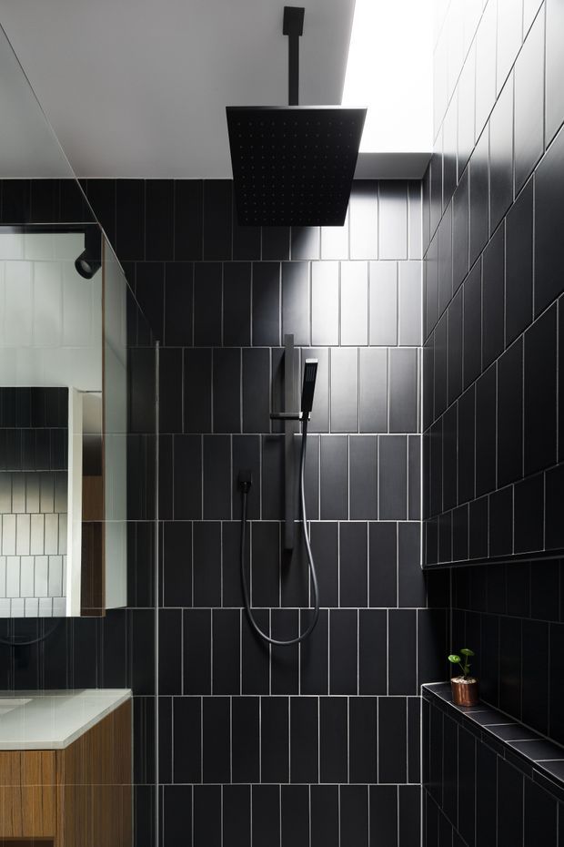 A compact contemporary bathroom shower area with floor-to-ceiling black rectangular wall tiles arranged in a grid. The shower features a large square rainfall showerhead, handheld shower, and matte black fixtures. A glass partition and partial floating timber vanity with a pale stone benchtop are visible at left, while a recessed tiled shelf with a small plant sits at right. The space has strong directional lighting and a dark, refined, high-contrast mood.