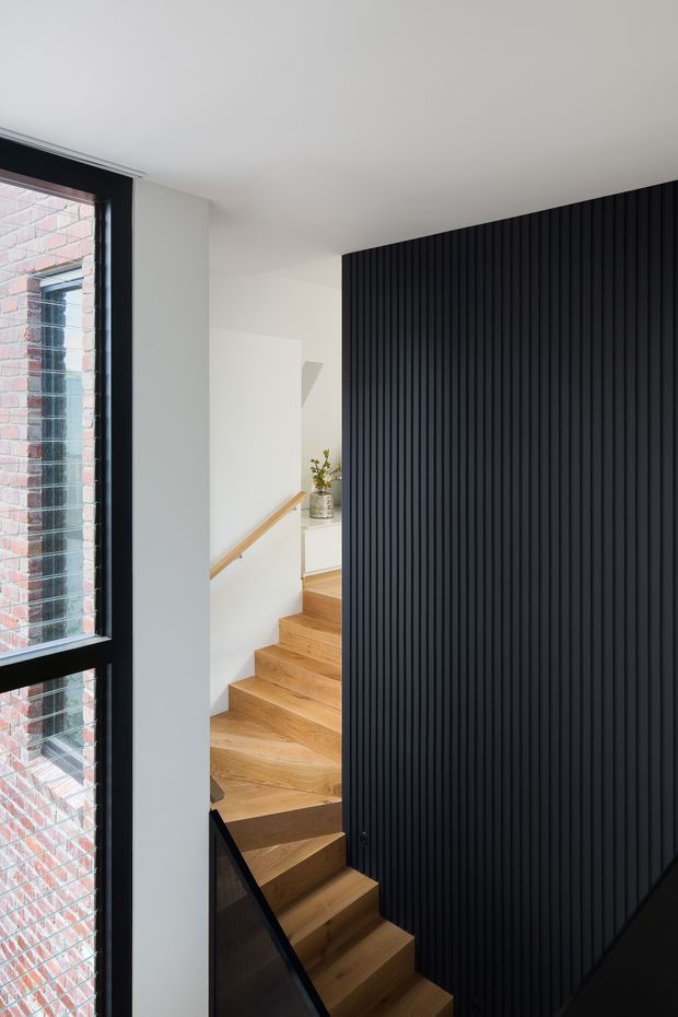 A modern stairway interior with warm timber stairs ascending through a compact landing, contrasted by a dramatic full-height black vertical-ribbed feature wall. A large black-framed window on the left reveals adjacent brickwork, while white walls and ceiling create a bright, minimalist composition. A timber handrail and small built-in landing console are visible.