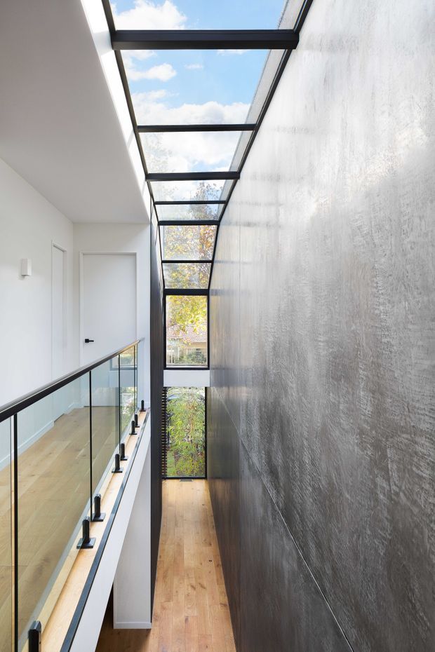 A narrow, bright interior circulation space viewed along an upper-level landing or hallway. A full-height textured grey feature wall runs along the right side, while a glazed skylight roof with dark metal framing extends overhead and admits abundant daylight. The left side has a clear glass balustrade with dark handrail overlooking a lower level, pale timber flooring, white walls, and simple white doors. Large windows at the far end provide views to trees and neighbouring roofs.
