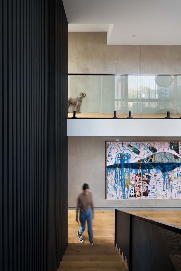 Interior architectural photograph looking down a stairway in a contemporary multi-level space. A dark vertically ribbed feature wall lines the left side, while warm timber stair treads and flooring contrast with pale textured wall surfaces. A glazed internal balustrade reveals an upper-level landing with a dog, and a large colourful abstract artwork is mounted on the wall below. Dark stair or balustrade joinery frames the foreground, with a person providing scale.