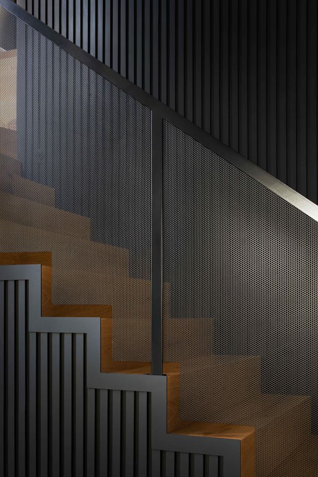 Close-up architectural detail of a contemporary stairway with warm timber treads, dark vertical slatted wall panelling and balustrade framing, and fine perforated metal mesh forming the stair guard. The composition is graphic and high-contrast, with predominantly black and charcoal surfaces contrasted by the wood.
