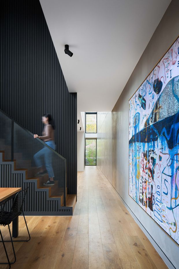 A contemporary residential hallway with a stairway along the left side. The stairs feature timber treads, a dark perforated or mesh balustrade, and a dramatic black vertical slatted wall. The hallway has wide pale timber flooring, white walls and ceiling, black track lighting, tall glazing at the far end, and a large colourful abstract artwork mounted on the right wall. A small portion of a timber dining table and black wire-frame chair is visible in the foreground.