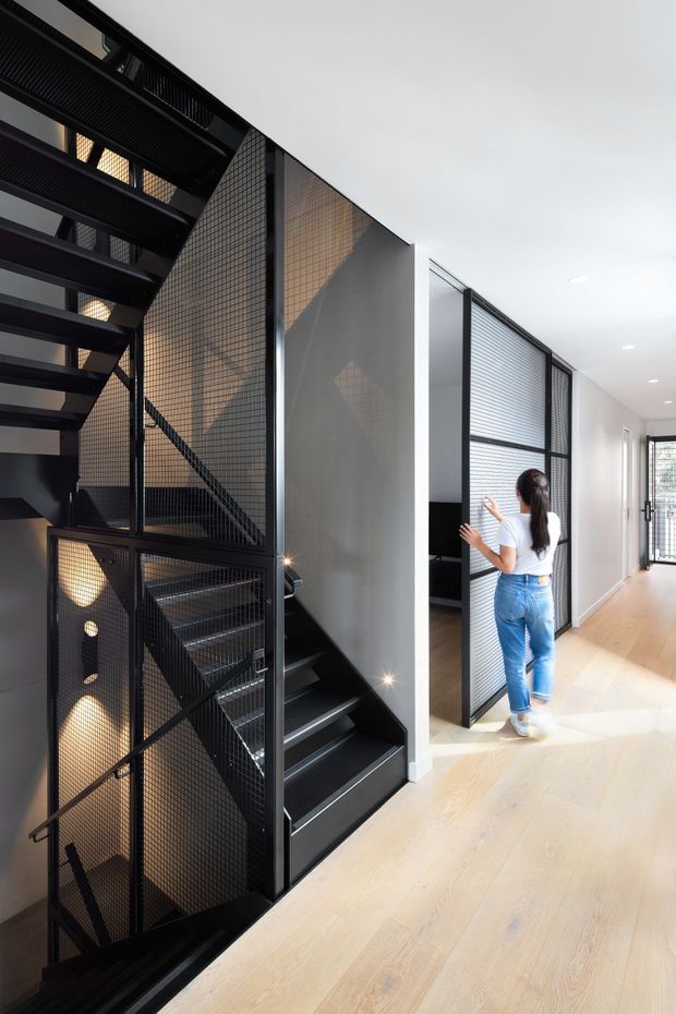 Bright contemporary interior hallway and stair landing featuring a dramatic black steel staircase enclosed with perforated metal mesh balustrades and screens. Pale timber flooring, white walls and ceilings, black-framed glazed and mesh doors, and a long corridor create a clean, modern residential setting.