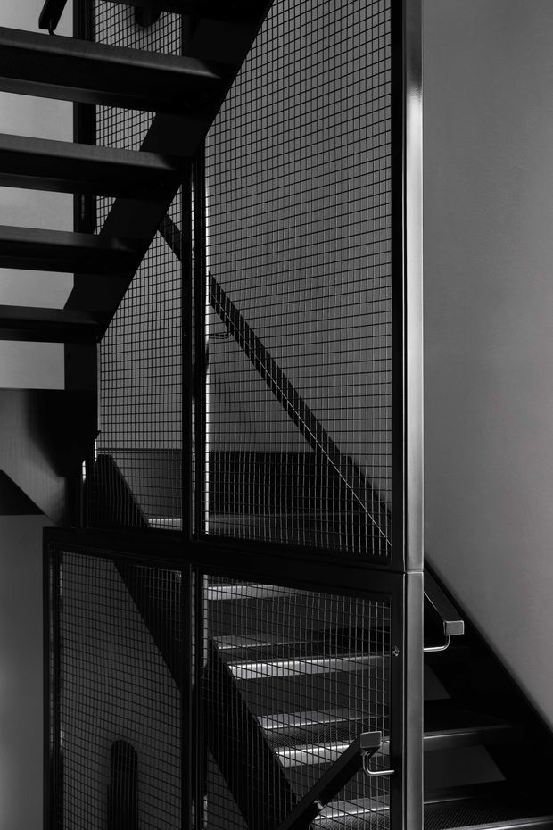A monochrome close-up architectural photograph of an enclosed industrial-style stairway. Black metal stair treads, structural stringers, handrails, and fine wire-mesh guards create strong geometric lines and layered shadows against pale grey walls.