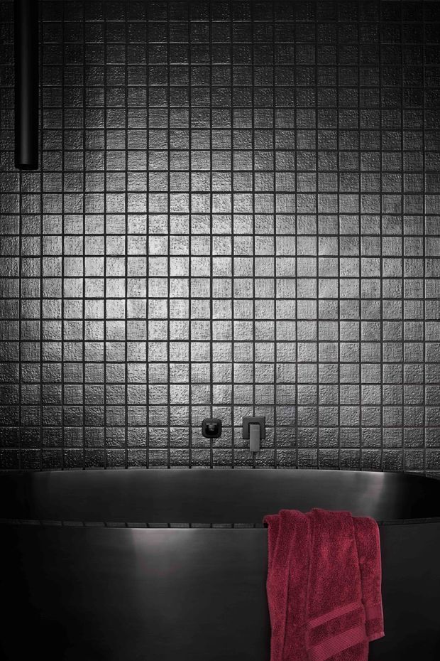 Moody close-up of a dark freestanding bathtub against a wall of small, textured square tiles. Minimal black tapware and fixtures are set into the tiled wall, while a deep burgundy towel provides the main colour accent.