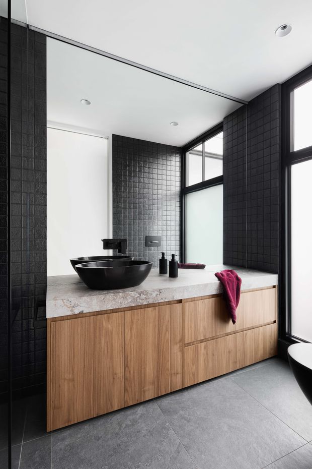Contemporary bathroom with a floating light-wood vanity, dark vessel basins and black wall-mounted tapware, a pale stone benchtop, charcoal square wall tiles, and large black-framed windows. A freestanding bath is partially visible at right. The room has a clean, high-contrast scheme with abundant natural light.