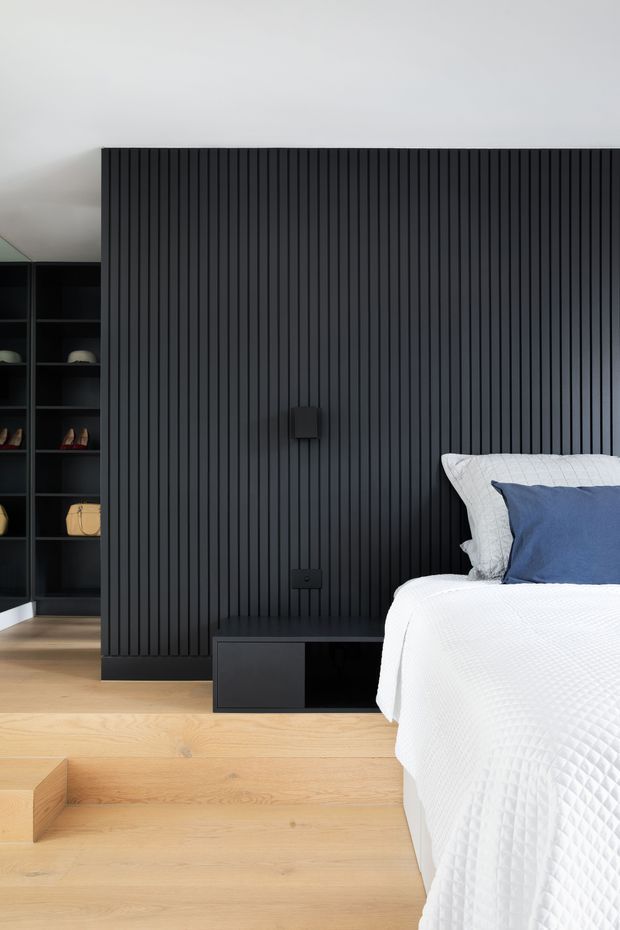 A contemporary bedroom featuring a dramatic full-height black vertical slatted feature wall, integrated black shelving and low cabinetry, pale timber flooring, and a partially visible bed dressed in white textured bedding with a blue cushion. The room has a clean, high-contrast, refined appearance.