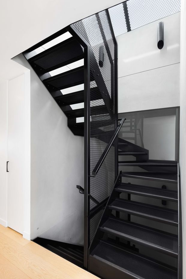 Interior view of a modern stairway with open-riser black metal stairs, perforated metal treads, and a black mesh balustrade. The staircase rises through a compact double-height void, contrasting with smooth white walls, a white built-in door, and pale timber flooring. Minimal black wall lights and strong geometric lines create an industrial, contemporary character.