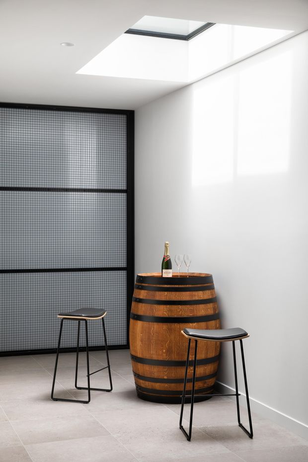 A bright, minimalist interior corner featuring a large timber wine barrel used as a bar table, with two slim black metal stools. A black-framed fine-grid glazed or screened partition occupies the left side, while white walls and ceiling are illuminated by a large skylight. Large pale floor tiles create a clean, contemporary setting.