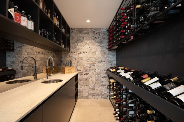 A compact, dark-toned wine room with extensive wall-mounted and rack storage for wine bottles. The space includes a long light stone benchtop with two sinks and brushed metal mixer taps, dark fixed cabinetry and open shelving, a textured printed feature wall resembling vintage wine labels or newspaper, and warm recessed ceiling lighting. Wine bottles and accessories fill the shelving, creating a functional cellar and tasting-preparation area.