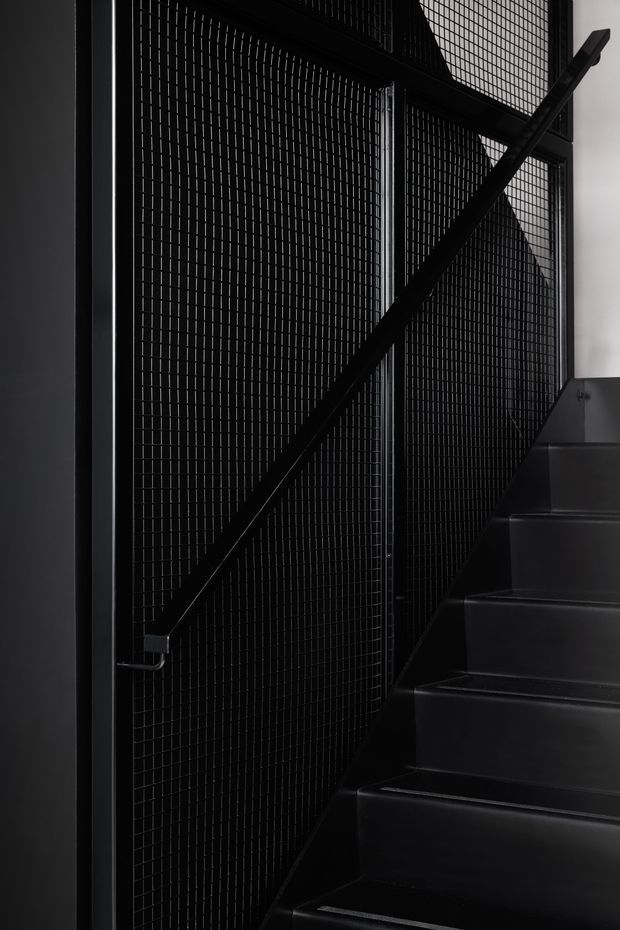 A dramatic close-up of a dark stairway with black metal stair treads, a black diagonal handrail, and a tall black expanded or wire-mesh screen forming the stair balustrade. The composition is highly graphic, monochromatic, and low-lit, with a small area of white wall visible at the upper right.
