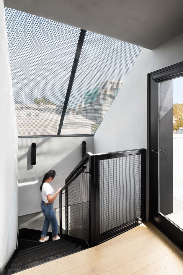 Bright contemporary interior stairwell with a descending black metal staircase and perforated mesh balustrade. White walls and angled glazing create an airy, light-filled space, while pale timber flooring contrasts with the dark stair and door frames. A large patterned translucent roof or skylight panel admits daylight and provides views toward neighbouring buildings.