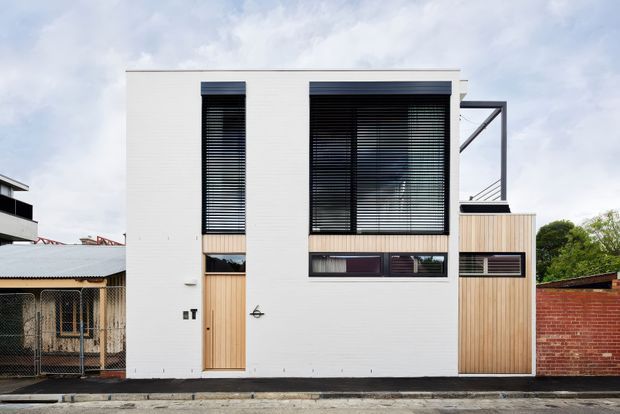 Street-facing view of a narrow contemporary two-storey residence with a crisp white rendered facade, tall black-framed windows fitted with dark horizontal blinds, and warm vertical timber cladding around the entry and side section. A simple steel roof or terrace frame projects above the right side, with neighbouring low-rise buildings and a brick wall visible around the property.