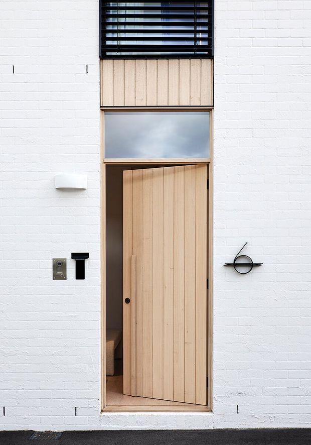 Close-up exterior view of a narrow modern entry set into a white-painted brick facade. The tall timber door has vertical slatted detailing and a long integrated pull, with a frosted transom window above and black horizontal louvres at the upper level. Minimal black wall-mounted lighting, intercom hardware, and a circular door fixture complete the restrained contemporary composition.