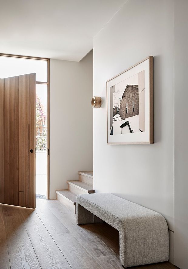 A refined residential entryway with a tall timber pivot door, pale timber flooring, and a short flight of stairs leading upward. A sculptural upholstered bench sits along the light neutral wall beneath framed monochrome artwork, with a small wall light and daylight entering through the glazed doorway. The space is calm, minimal, and softly contemporary.