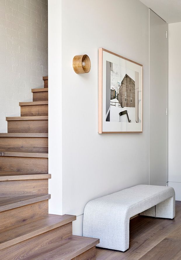 A bright, refined interior entry area with a floating timber staircase along a textured white-painted brick wall. A pale upholstered curved bench sits beneath a framed architectural print, with a small timber wall sconce and concealed light-grey cabinetry or a door to the right. Warm timber flooring and stair treads contrast with the white and soft neutral surfaces.