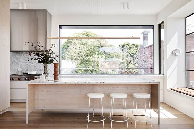 Bright contemporary kitchen with a large pale timber island and white stone benchtop, three white bar stools, white cabinetry, and a marble-look splashback. A wide black-framed window provides leafy views, while a slim linear pendant and clerestory-style glazing enhance the airy space. Warm timber flooring and a white panelled ceiling add subtle texture.