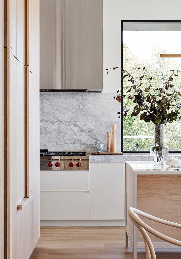 A refined contemporary kitchen featuring pale timber cabinetry, white cabinetry, a veined marble splashback and benchtop, integrated stainless-steel cooking appliances, and light timber flooring. A large black-framed window brings in natural light and overlooks greenery. The foreground includes a timber island and curved timber dining chairs.