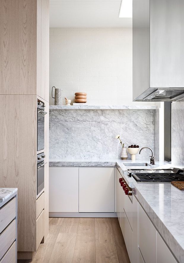 A bright, narrow contemporary kitchen with full-height pale timber cabinetry, integrated stainless-steel ovens, a marble-look stone splashback and benchtops, white lower cabinetry, a white painted brick wall, timber flooring, and a large stainless-steel rangehood. A skylight brings natural light into the compact space, with a sink and gas cooktop along the right-hand counter.