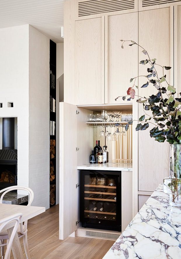 A bright, refined interior showing a built-in light timber bar and wine storage cabinet. The open cabinet contains hanging stemware, bottles, and a compact glass-front wine fridge, with marble benchtops extending through the foreground. A white brick fireplace and integrated firewood storage are visible to the left, along with part of a dining area. The room has pale timber floors, light vertical cabinetry, and a slatted ceiling.
