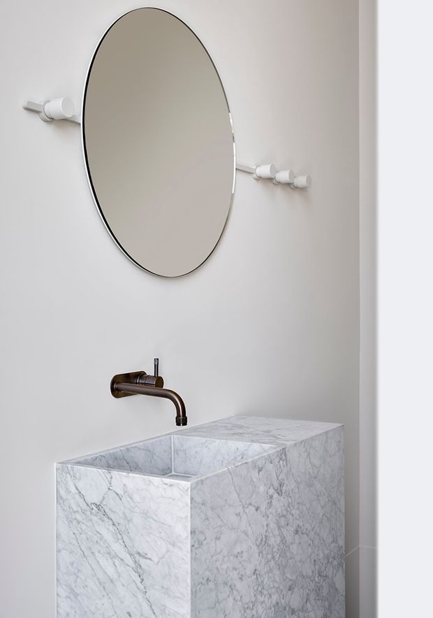 A minimalist bathroom vanity featuring a sculptural white-grey veined marble basin and countertop with an integrated rectangular sink. A dark bronze wall-mounted faucet sits beneath a large oval mirror, accompanied by a row of white cylindrical wall hooks or fittings. The space has plain white walls and a clean, refined contemporary appearance.