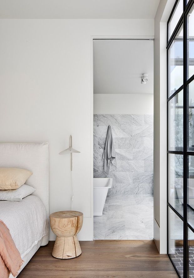 Bright, minimalist bedroom with a partially visible upholstered bed, layered neutral bedding, a small sculptural timber stool, and a wall-mounted pendant. A bathroom opens directly from the bedroom, featuring pale grey-white marble-look wall and floor tiles, a toilet, and a towel. Black-framed glazed doors or windows line the right side, bringing in abundant natural light. The room has a calm, refined neutral palette and warm timber flooring.
