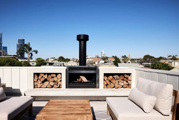A bright rooftop terrace with a built-in outdoor fireplace and chimney centred in a rendered white firewood surround. Split firewood is stored in open niches on both sides, while cushioned outdoor sofas and a timber coffee table create a relaxed seating area. A low white parapet frames a clear urban skyline in the background.