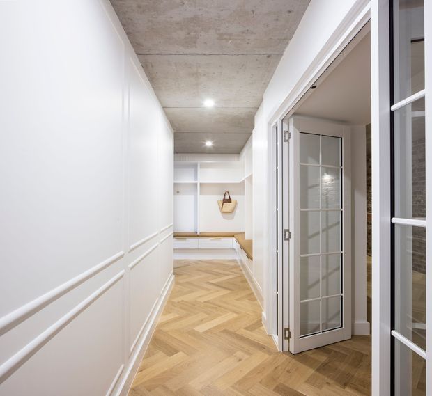 A bright, refined hallway with white panelled walls, a raw exposed concrete ceiling, and warm timber herringbone flooring. Built-in white cabinetry and a bench occupy the far end, while glazed internal doors open to adjacent rooms on the right.