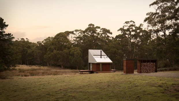 A small minimalist cabin set in a grassy clearing at the edge of dense eucalyptus woodland. The compact structure has a steep pitched roof with a large glazed roof plane or skylight, dark timber cladding, and a simple deck. A separate timber outbuilding with a stacked firewood store sits beside it. The scene is photographed in warm, muted evening light and has a secluded rural character.