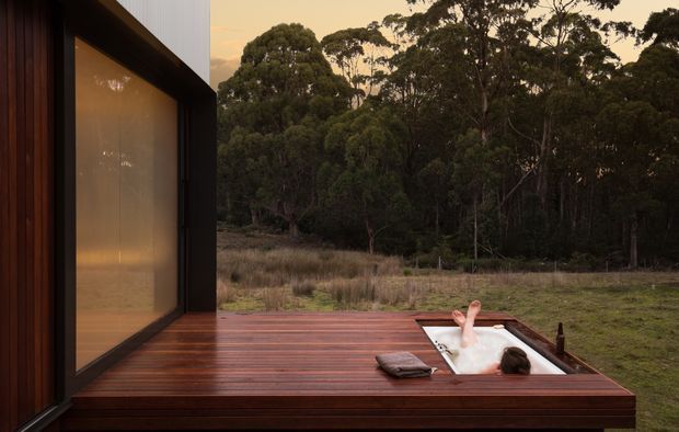 A dusk exterior view of a modern house opening onto a dark timber deck. The deck contains a recessed freestanding white bathtub with a person bathing, while warm light glows through the large glazed wall. Dense native woodland and grassland form the surrounding landscape.