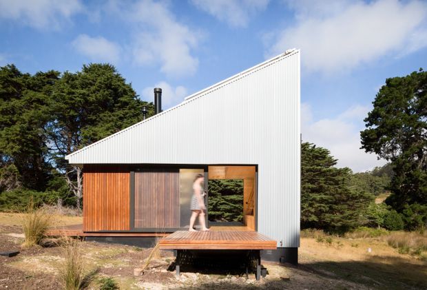 Exterior view of a compact contemporary single-storey dwelling set in a natural, wooded landscape. The building has a sharply sloping mono-pitched roof clad in white ribbed metal, warm vertical timber wall cladding, dark-framed glazing, and a raised timber deck projecting from the front. A person is visible near the glazed opening, with mature trees and dry grasses surrounding the structure.