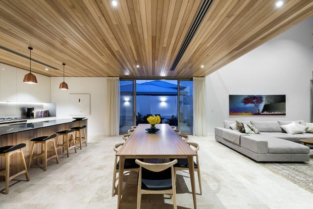 Open-plan contemporary kitchen, dining and living area with a long timber dining table, kitchen island and stools, large sectional sofa, and full-height glazing at the rear. The space features a striking linear timber ceiling, recessed lighting, pale flooring, white walls and warm wood joinery.