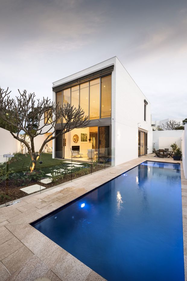 A contemporary two-storey white rendered house with a strong rectangular form, expansive floor-to-ceiling glazing, and a narrow swimming pool extending along the side. The pool terrace is paved in light stone, with a landscaped courtyard garden, sculptural pruned trees, glass fencing, and outdoor dining visible near the rear. Warm interior lighting contrasts with the cool dusk exterior.