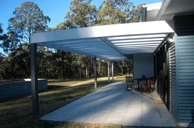 A contemporary covered outdoor patio beside a corrugated metal-clad building, with a broad concrete-paved floor, slim dark steel posts, and a translucent or open slatted roof canopy. A small outdoor dining setting sits along the wall, while a low masonry retaining wall and wooded landscape are visible beyond.