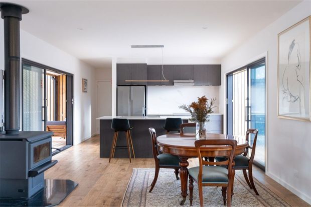 Bright open-plan kitchen and dining area with warm timber flooring, dark built-in kitchen cabinetry, a central island with stools, and a traditional wooden dining table and chairs. Large glazed doors and windows bring in abundant daylight, with a freestanding wood-burning stove visible at left and framed artwork on the wall.